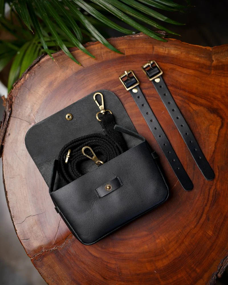 Geöffnete schwarze Trip Machine Sidekick Moto Pouch mit Schulterband und zwei Befestigungsriemen auf einer Holzscheibe.