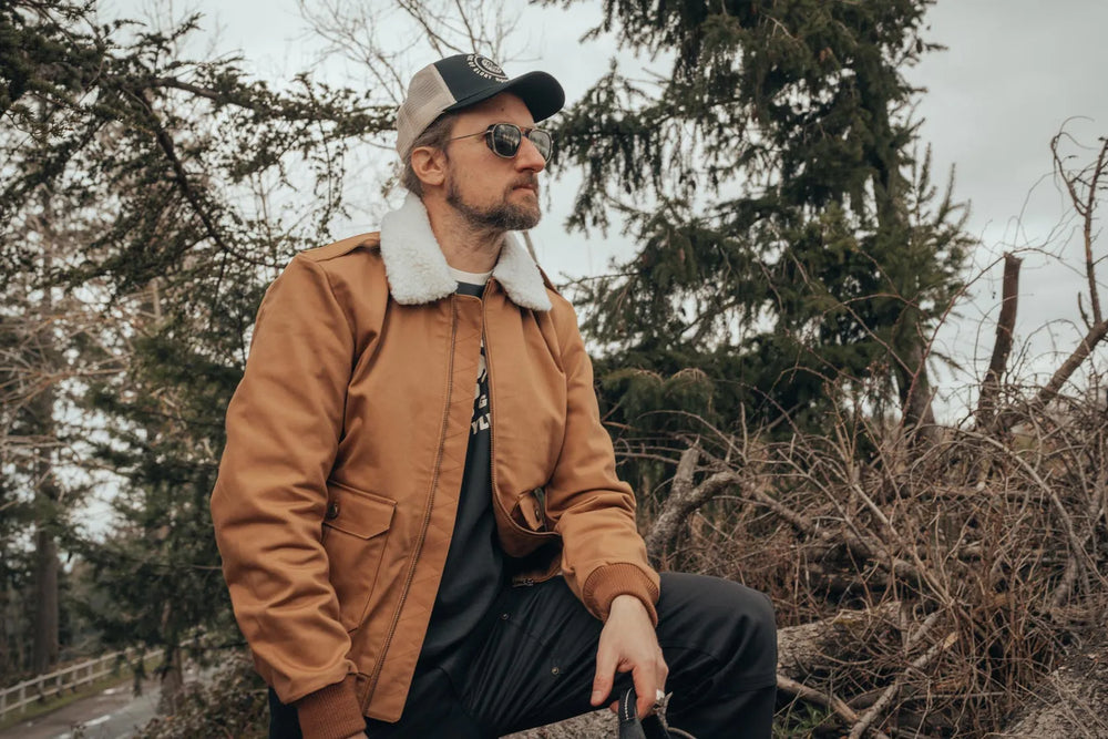Mann mit Cap und Sonnenbrille trägt die karamellfarbene Age of Glory Cockpit Jacke mit hellem Kragen im Wald.