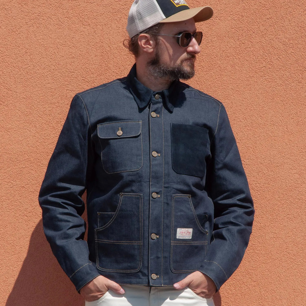 Mann mit Cap und Sonnenbrille trägt die Age of Glory Craftsman Denim Jacke vor einer orangefarbenen Wand.